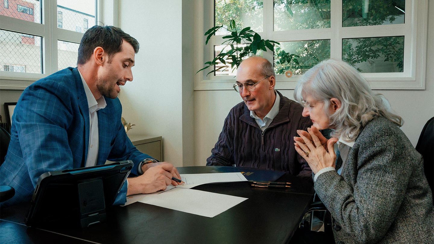 Alpha Bull Canada financial advisor explaining investment options to a senior couple in an office setting. Advisor in a blue suit presenting documents while elderly clients attentively listen, showcasing a professional financial consultation environment.